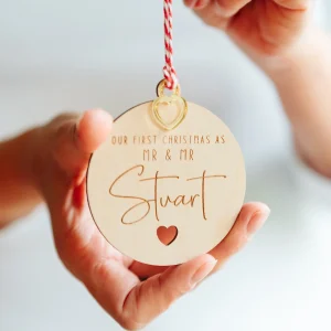 Home 5 Hand holding a personalised wooden Our First Christmas as Mr & Mr bauble engraved Stuart with a gold heart charm